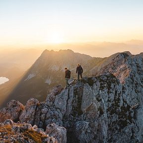 Zwei Wanderer stehen bei Sonnenaufgang auf einem felsigen Berggipfel. Im Hintergrund erheben sich weitere Berge und ein See ist in der Ferne sichtbar.