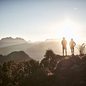 Two hikers stand silhouetted against a sunset backdrop over the Wilder Kaiser mountains, with rolling hills and dramatic peaks visible in the soft evening light.