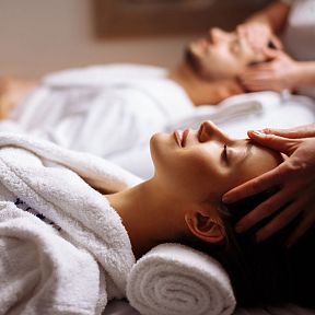 Zwei Personen in weißen Bademänteln genießen eine entspannende Kopfmassage in einem Spa. Sie liegen auf weichen Handtüchern und wirken entspannt und erholt.