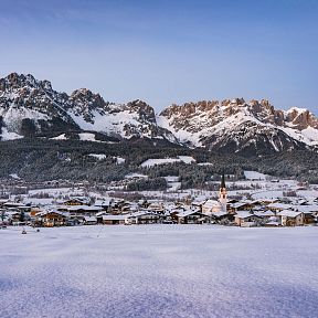 A picturesque winter scene with a snow-covered village below the rugged peaks of the Wilder Kaiser mountains, under a clear blue sky.
