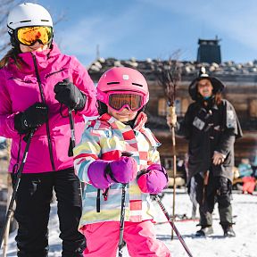 A family enjoying skiing at Wilder Kaiser. The scene features a child in colorful ski gear, adults in winter attire, and a traditional mountain hut in the background.