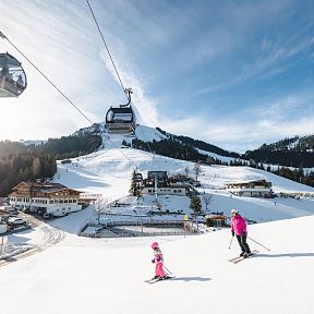 Winterliche Skiszene in Tirol mit Bergkulisse, Skifahrern und einer Seilbahn. Schnee bedeckt die Landschaft, während sich Skifahrer die Piste hinabbewegen.