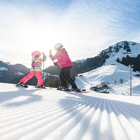Ein Kind und ein Erwachsener in bunter Skikleidung fahren bei Sonnenschein auf einer schneebedeckten Piste in den Bergen.
