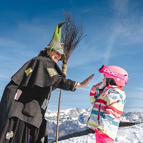 Eine Person in einem Hexenkostüm mit Besen klatscht in die Hand eines Kindes mit pinkem Helm. Hintergrund: schneebedeckte Berge und blauer Himmel.