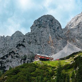 Eine Berghütte liegt auf einem grünen Hügel vor einer beeindruckenden Felsenkulisse unter blauem Himmel. Tannenbäume stehen im Vordergrund.