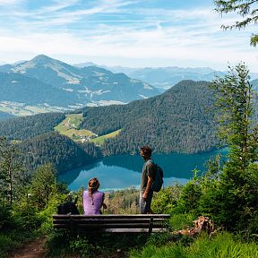 Zwei Wanderer sitzen auf einer Bank und blicken auf einen blauen Bergsee umgeben von grünen Hügeln und Wäldern in einer idyllischen Berglandschaft.