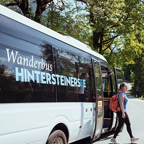 Eine Frau mit Rucksack steigt aus einem Wanderbus mit der Aufschrift "Hintersteinersee". Im Hintergrund sind Bäume und ein sonniger Himmel zu sehen.