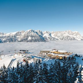 Winter view of the snow-covered Wilder Kaiser mountains with a ski lodge surrounded by pine trees and clear blue sky in the background.