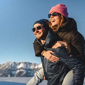 Ein lachendes Paar mit Winterkleidung genießt einen sonnigen Tag in schneebedeckten Bergen. Sie tragen Sonnenbrillen und warme Mützen.