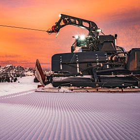 A snow grooming machine prepares ski slopes at sunset in Wilder Kaiser, with orange skies and mountains in the background. The snow is freshly groomed.