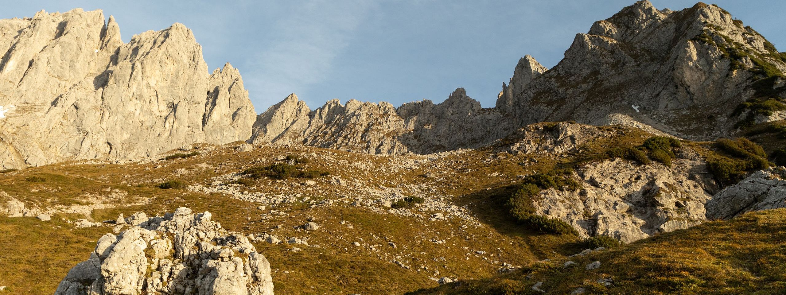 A golden-hued view of the Wilder Kaiser mountains with rocky peaks and gentle grassy slopes under the warm light of sunset, creating a picturesque alpine scene.