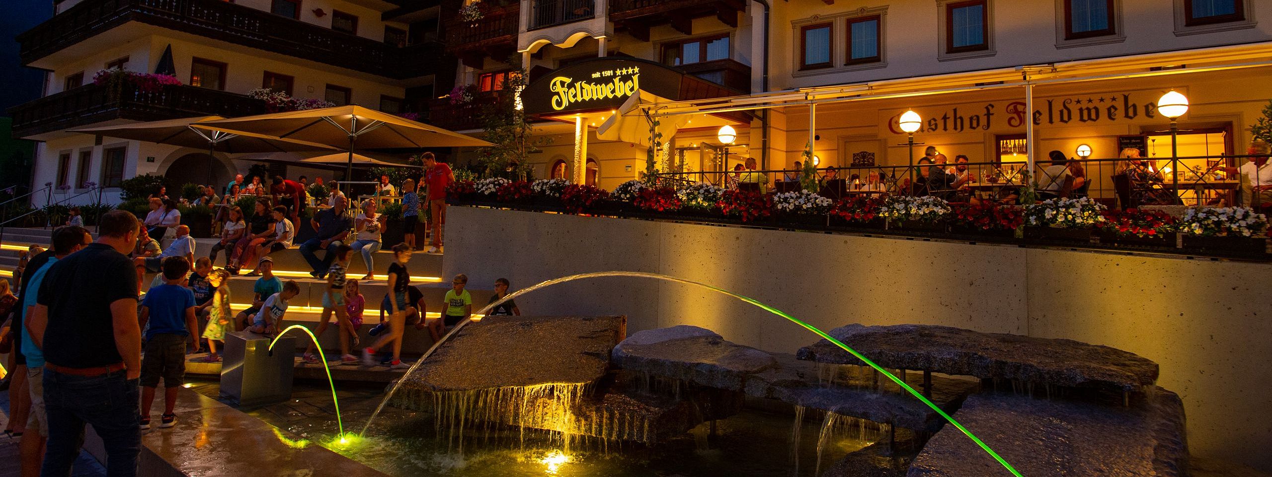 Eine belebte Terrasse eines alpenländischen Hotels bei Abenddämmerung. Menschen entspannen, und Lichter erleuchten die Fassade, während ein Brunnen im Vordergrund sprudelt.