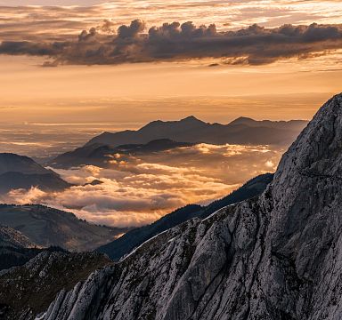 Ein beeindruckendes Gebirgspanorama mit felsigen Gipfeln im Vordergrund, umgeben von Wolken und einem orangefarbenen Sonnenuntergangshimmel in der Ferne.