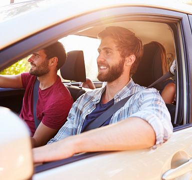 Ein Auto fährt eine Landstraße entlang, umgeben von grünen Wiesen und Bäumen. Im Hintergrund sind schneebedeckte Berge unter einem blauem Himmel zu sehen.