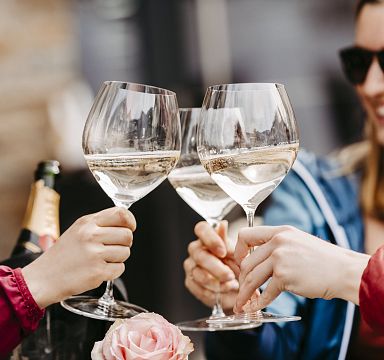 Personen stoßen mit Weißweingläsern an, im Hintergrund unscharf eine Flasche Sekt. Ein rosa Blumenarrangement schmückt den Tisch.