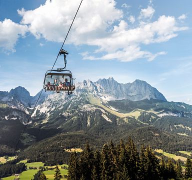 Eine Seilbahn schwebt über grüne Wälder und Wiesen, im Hintergrund erhebt sich majestätisch das Bergmassiv des Wilden Kaisers unter einem blauen Himmel mit Wolken.