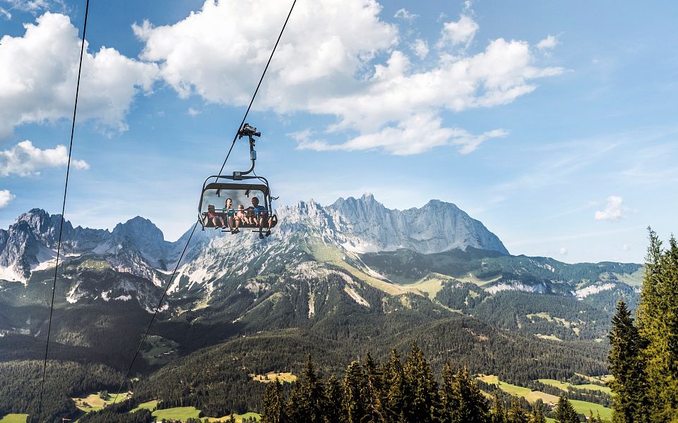 Eine Seilbahn schwebt über grüne Wälder und Wiesen, im Hintergrund erhebt sich majestätisch das Bergmassiv des Wilden Kaisers unter einem blauen Himmel mit Wolken.