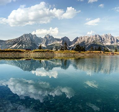 Bergpanorama des Wilden Kaisers spiegelt sich in einem klaren, ruhigen See unter einem blauen Himmel mit weißen Wolken. Grüne Wälder säumen das Ufer.