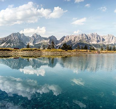 Ein klarer Bergsee spiegelt die dramatischen Gipfel des Wilden Kaisers unter einem blauen Himmel mit weißen Wolken. Die Szenerie wirkt friedlich und majestätisch.