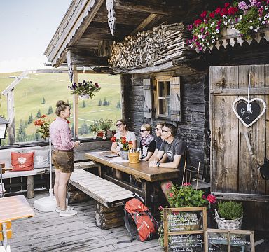 Gruppe von Menschen sitzt an einem Holztisch vor einer rustikalen Berghütte, während eine Frau in traditioneller Kleidung mit ihnen spricht. Blumen schmücken die Hütte.