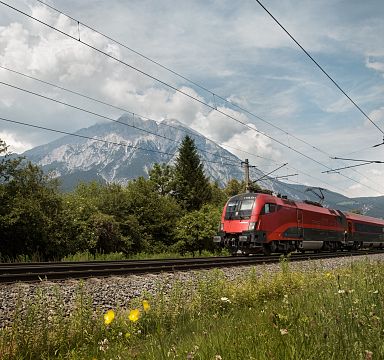 Roter Zug fährt durch grüne Landschaft, im Hintergrund beeindruckendes Bergmassiv unter blauem Himmel mit Wolken.