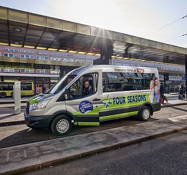 Ein grüner Shuttlebus mit der Aufschrift "Four Seasons" steht auf einer städtischen Straße, im Hintergrund sind moderne Gebäude und eine Bushaltestelle zu sehen.