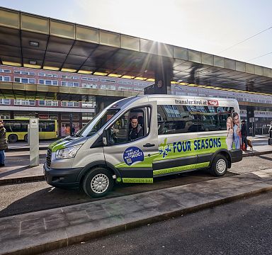 Ein grüner Shuttlebus mit der Aufschrift "Four Seasons" steht auf einer städtischen Straße, im Hintergrund sind moderne Gebäude und eine Bushaltestelle zu sehen.