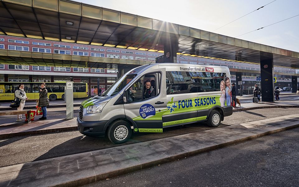 A branded shuttle bus labeled "Four Seasons" parked at a busy transportation hub, with modern buildings and passengers in the background.