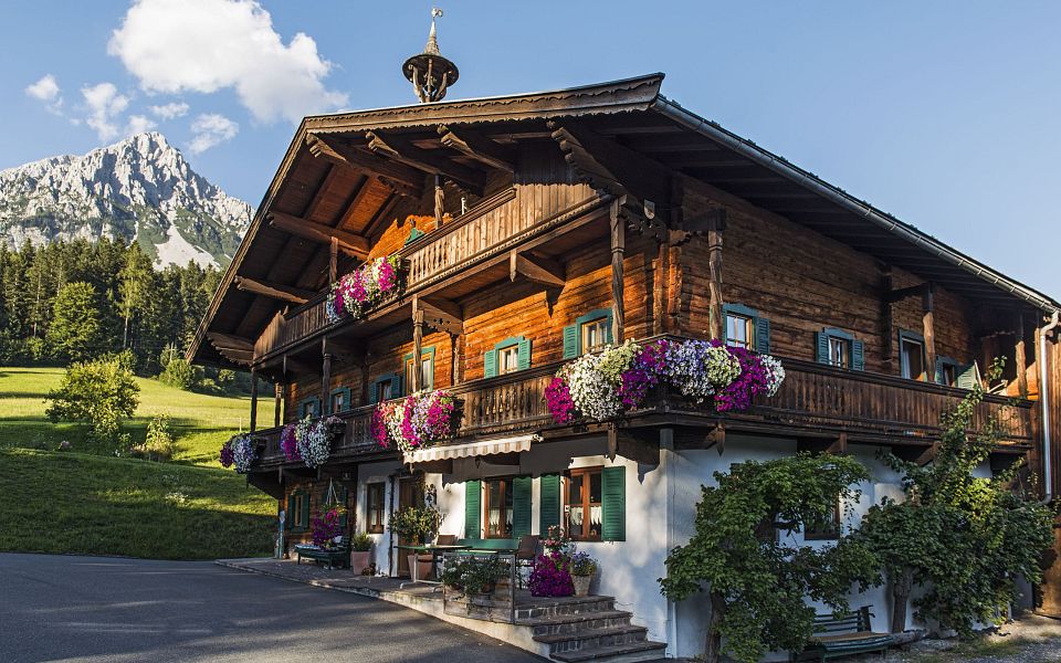 Ein traditionelles Tiroler Holzhaus mit üppigen Blumenbalkonen vor einem grünen Hügel und einem majestätischen Bergmassiv im Hintergrund.