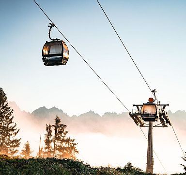 Cable cars ascending with the majestic Wilder Kaiser mountains in the background, surrounded by trees and a hazy sky at sunrise.