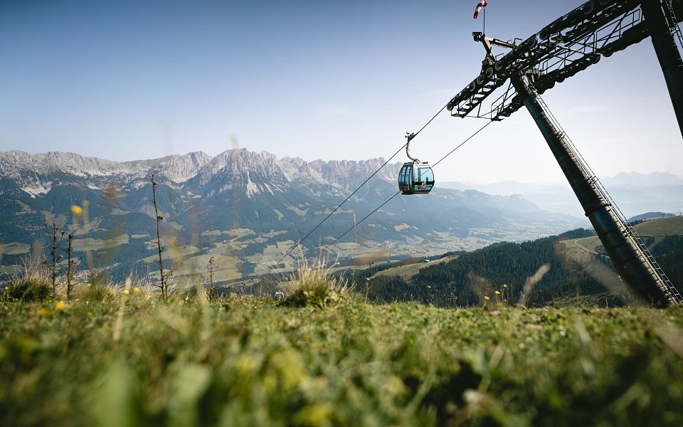 Blick über eine grüne Bergwiese auf eine Seilbahn mit Kabine vor einem Panorama von majestätischen Bergen unter klarem, blauem Himmel.
