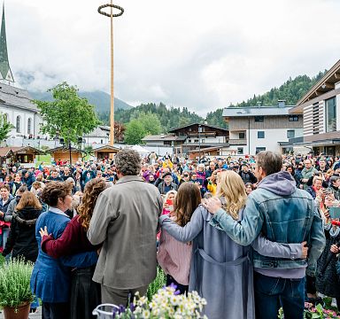Eine große Menschenmenge auf einem Dorfplatz, umgeben von Bergen. Im Vordergrund umarmen sich mehrere Personen und schauen in die Menge.