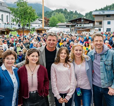 A group of people posing for a photo at an outdoor event, with a large crowd and mountain scenery in the background.