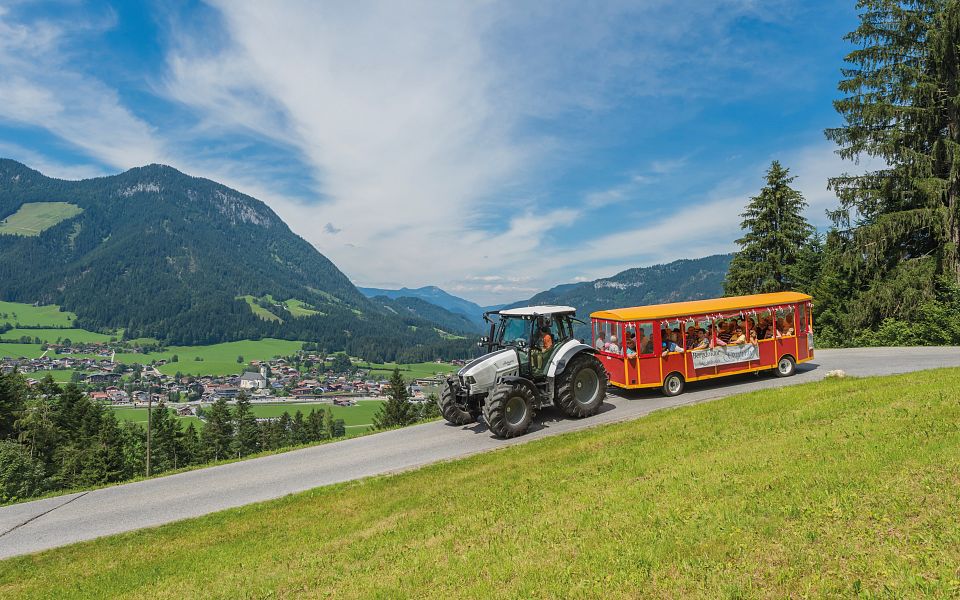 Ein Traktor zieht eine rote Touristenbahn den Hügel hinauf, umgeben von grüner Landschaft und Bergen im Hintergrund.