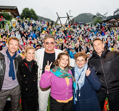 A large crowd in raincoats at an event in the Wilder Kaiser region, standing outside with mountains in the background. Six people in front posing, making peace signs.