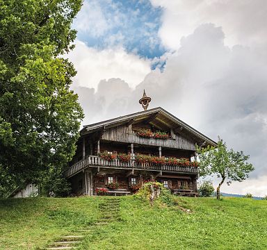 Holzhütte in Berglandschaft mit blauem Himmel, großen Bäumen und grüner Wiese. Traditionelle Architektur mit Blumen geschmücktem Balkon.