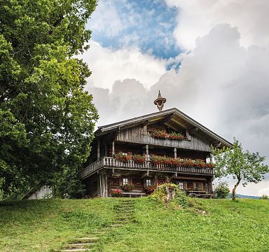 Holzhütte in Berglandschaft mit blauem Himmel, großen Bäumen und grüner Wiese. Traditionelle Architektur mit Blumen geschmücktem Balkon.