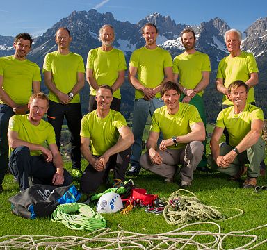 Eine Gruppe von Männern in grünen T-Shirts posiert mit Kletterausrüstung auf einer Wiese. Im Hintergrund sind beeindruckende Berge zu sehen.