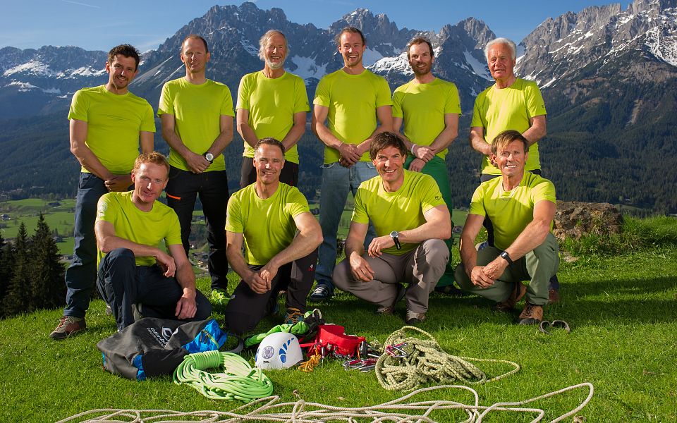 Eine Gruppe von Männern in grünen T-Shirts posiert mit Kletterausrüstung auf einer Wiese. Im Hintergrund sind beeindruckende Berge zu sehen.