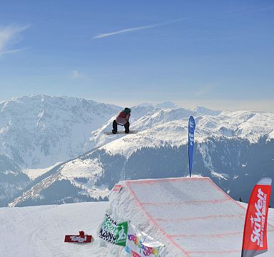 Snowboarder in der Luft über einer verschneiten Rampe mit Alpen im Hintergrund. Klare blaue Himmel und schneebedeckte Berge bieten eine beeindruckende Kulisse.