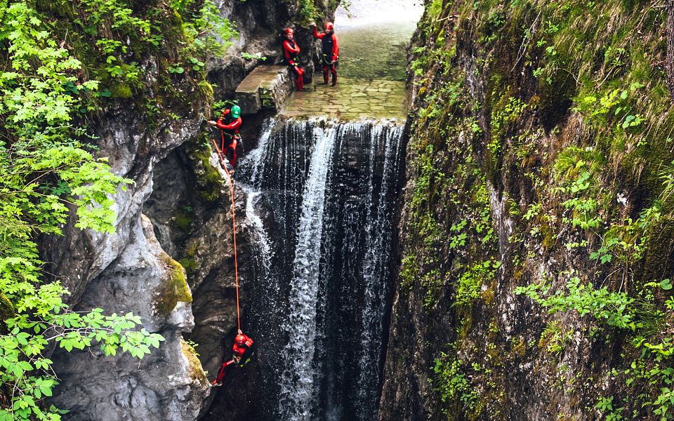 Zwei Personen seilen sich an einem Wasserfall in einer felsigen, bewaldeten Schlucht ab. Die Szene ist dynamisch und von dichtem Grün umgeben.