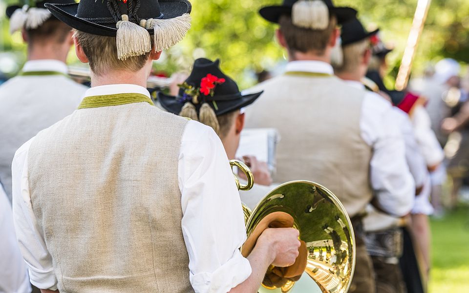 Blasmusikanten in traditioneller Tracht mit Hüten und Instrumenten, von hinten fotografiert, spielen im Freien bei sonnigem Wetter in einer grünen Umgebung.