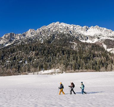 A couple walking in the snow with the Wilder Kaiser mountains in the background under clear blue skies.