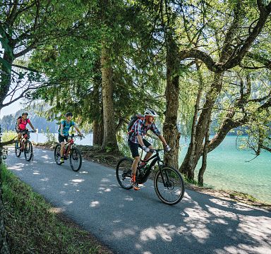 Cyclists on a path near a scenic lake surrounded by trees in the Wilder Kaiser region.