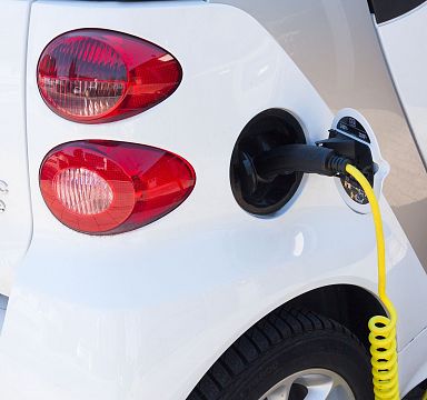 Ein weißes Elektroauto wird mit einem gelben Ladekabel an eine Ladestation angeschlossen. Im Hintergrund sind Rücklichter zu sehen. Nahaufnahme.