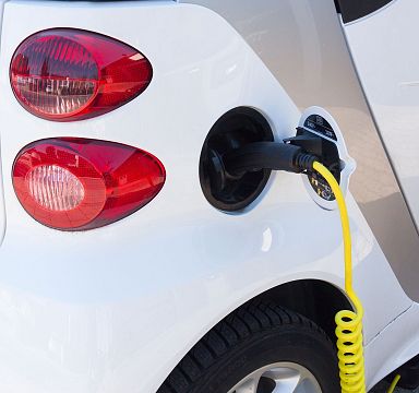 Ein weißes Elektroauto wird mit einem gelben Ladekabel an eine Ladestation angeschlossen. Im Hintergrund sind Rücklichter zu sehen. Nahaufnahme.