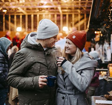 Ein lächelndes Paar in Winterkleidung steht auf einem belebten Weihnachtsmarkt, umgeben von Lichtern und stimmungsvoller Dekoration. Es schneit leicht.