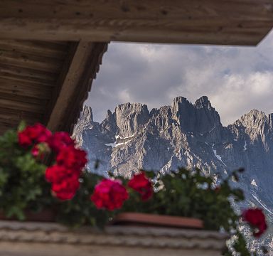 A scenic view of the Wilder Kaiser mountains framed by red flowers, capturing the majestic peaks and a touch of traditional Alpine architecture.