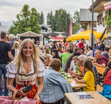 Eine fröhliche Frau in traditioneller Tracht steht vor einem belebten Biergarten mit vielen Menschen an Tischen im Freien bei sommerlichem Wetter.