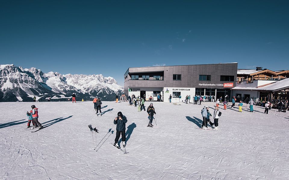 Skifahrer auf einer verschneiten Bergstation mit modernem Gebäude. Im Hintergrund beeindruckendes Alpenpanorama unter klarem, blauem Himmel.